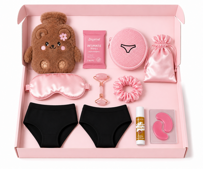 A curated self-care gift set arranged neatly inside a light pink box. The set includes a brown plush teddy bear, a pink satin sleep mask, black underwear, a pink round pouch, a pink drawstring bag, a pink jade roller, a pink scrunchie, intimate wipes, and a bottle of lip balm.