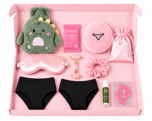 A curated self-care gift set arranged neatly inside a pink box on a white background. The set includes black underwear, a pink satin sleep mask, a pink drawstring pouch, a green plush dinosaur toy, a pink round pouch, a pink packet of intimate wipes, a rose-gold facial roller, a pink scrunchie, and other small wellness items.